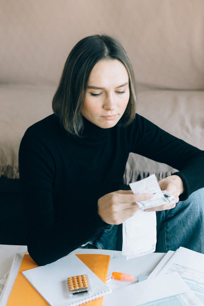 Young woman in black sweater reviewing bills at home, using a calculator, surrounded by documents.